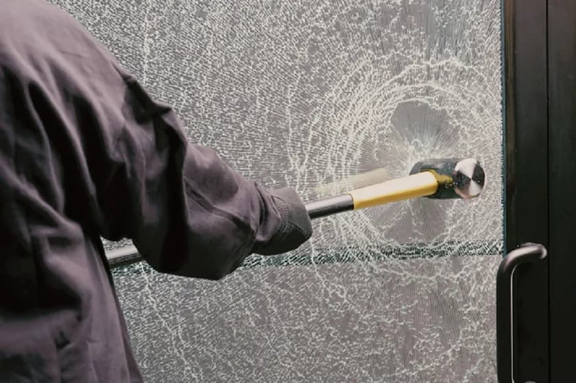 A person in a dark hooded jacket swinging a sledgehammer into a commercial glass door; the pane has crazed white across its full width but has not opened