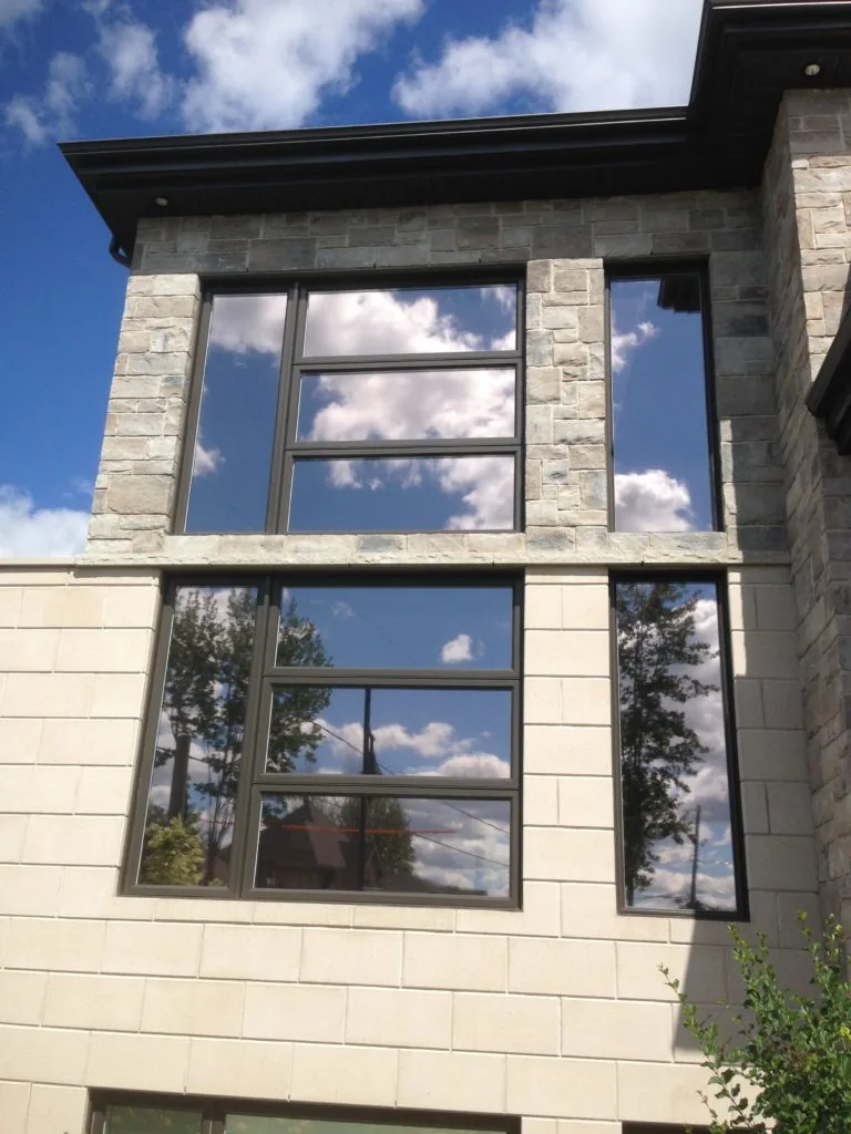 Stone-clad Edmonton home with tall windows tinted by XPEL VISION film, mirroring a bright sky